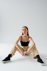 full length of pretty model in beige pants and black top looking at camera while sitting on grey.