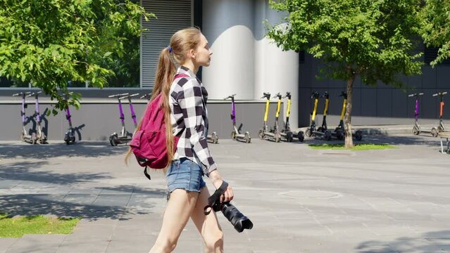 Cute girl with professional camera walks along paved path in summer in city. Side view. Photographer traveler with a backpack. Female student photographer goes on walkway, surrounded by