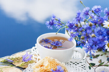 Obraz na płótnie Canvas Cornflower herbal tea in white cup on white crochet napkin on wooden table outdoors, healthy cornflower drink with honey, fresh cornflowers flowers in vase, healthcare and healthy eating concept