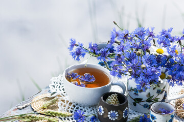 Obraz na płótnie Canvas Cornflower herbal tea in white cup on white crochet napkin on wooden table outdoors, healthy cornflower drink with honey, fresh cornflowers flowers in vase, healthcare and healthy eating concept