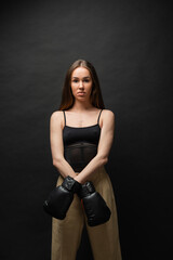 brunette woman in top and beige pants posing in boxing gloves on black background.