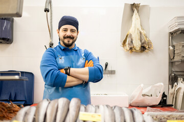 A smiling middle-aged fishmonger poses with his arms crossed behind the sales counter, selling...