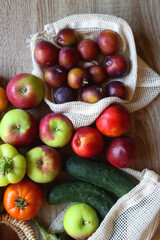 Straw bag and reusable fabric bags filled with various healthy fruit and vegetables. Wooden background, top view.