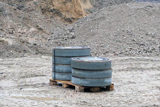 Konzept Baustelle: Eine Palette mit neuen Gullydeckeln, Regenwassereinlauf oder Sinkkasten, auf einem neuen Bauplatz mit viel Erdaushub im Hintergrund