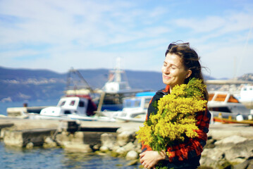 A pretty girl laughs, turning her face to the sea wind and standing with a large bunch of bright yellow mimosa flowers against the backdrop of ships in the port