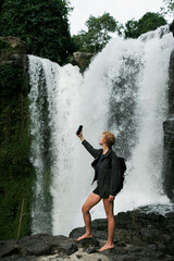 A young beautiful and cheerful girl takes a selfie on the phone against the backdrop of a waterfall.