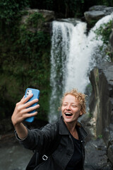 Fototapeta premium A young beautiful and cheerful girl takes a selfie on the phone against the backdrop of a waterfall.