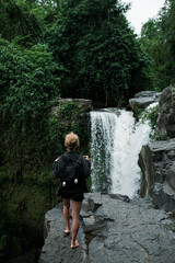 A young blonde girl with a backpack travels to a large waterfall, rear view.