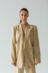 brunette young woman in beige jacket posing while looking at camera isolated on grey.