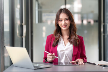 Pretty Asian businesswoman sitting smiling and happy working on laptop computer and document in office. Looking at camera.