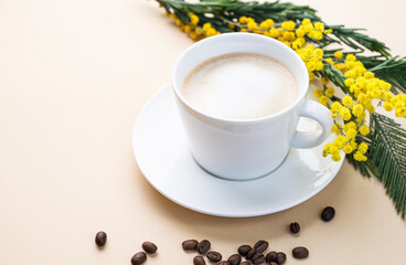 A spring bouquet with yellow mimosa flowers and a cup of coffee  cappuccino on a beige table with coffee beans. Concept of 8 March, happy women's day.