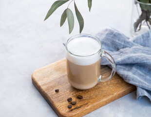 Cappuccino or latte with milk foam in glass on a wooden plate on a light blue background with coffee beans and eucalyptus branch. The concept of spring breakfast.