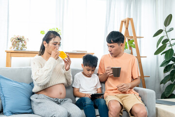 Asian father brings orange juice to a pregnant mother teaching her child homework on tablet. and man is holding a coffee cup in his hand while he is sitting in the middle.