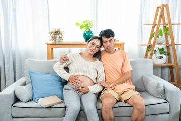Obraz premium Asian couple. Pregnant wife sitting and talking. Pregnant woman sitting next to her reading a book on the sofa in the house.