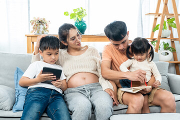 family love warmth Asian parents are relaxing on the sofa in living room of their home. their son and daughter are drawing on  coloring book with mom and dad guiding. Pregnant woman. Big belly.