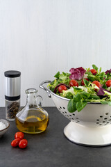 lettuce leaves and cherry tomatoes in a colander on a gray table, olive oil, salt and pepper. White background, copy space, front view