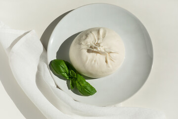 Traditional Italian, fresh, soft burrato ball of young mozzarella cheese with cream on a round white plate with basil greens on a light background. close-up
