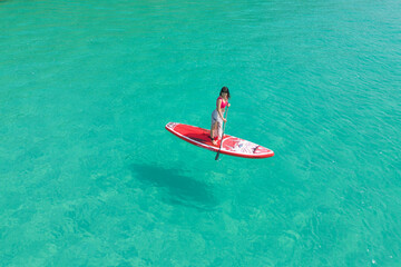 Fototapeta premium Aerial top view of Asian woman, a tourist, paddling a boat, canoe, kayak or surfboard with clear blue turquoise seawater, Andaman sea in Phuket island in summer season, Thailand. Water in ocean