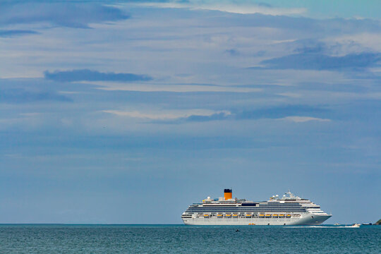 navio de cruzeiro navegando em &aacute;guas calmas no horizonte e uma pequena lancha passando ao seu lado em dia de sol, c&eacute;u azul e algumas nuvens.
