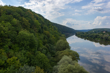 the river flows through the canyon. Hills with forests on one side, green fields on the other. A cloudy blue sky reflects in the river