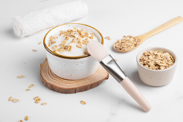 Homemade facial mask or creamy scrub with cereal, made of yogurt and oats flake, home spa cosmetics with brush on white marble background with natural beauty treatment ingredients and towel, close up