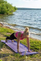 An elderly woman in sportswear standing in a plank, practicing yoga on the shore of the lake. Yoga in the park.