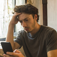 Young man looks at his cell phone worried and holds his head with one hand