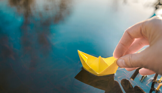 Yellow lonely floating paper boat in hand. Colorful sailing ship in big blue spring puddle, river water.Warm wet rainy weather.Hello spring,autumn.Children play,have street fun outdoors.Way to future