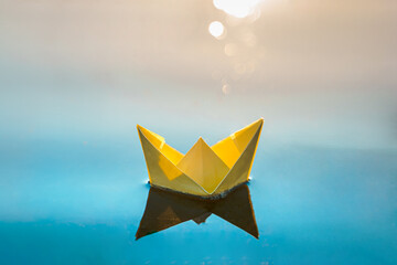 Fototapeta na wymiar Yellow floating paper boat. Colorful sailing ship in big blue spring puddle, river water on winter street. Warm wet rainy weather, old grass. Hello spring, autumn. Children play, have fun outdoors
