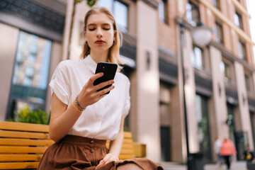 Low-angle view of pretty blonde young woman using typing mobile phone sitting on bench on city street on blurred background of modern building, thinking looking on device screen outdoors.