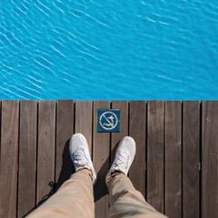 Man with beige trousers and fashion white sneakers stands on a wooden floor near a swimming pool with blue water. Do not dive vintage sign