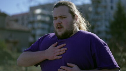One young overweight man having chest pains standing outdoors. A fat person feeling stressful pain...
