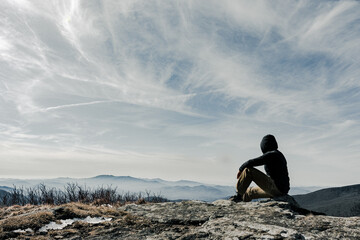 Male hiker sitting on top of the mountain in cold winter afternoon, looking at horizon. Man resting on the rock. Concept motivation and goal achievement
