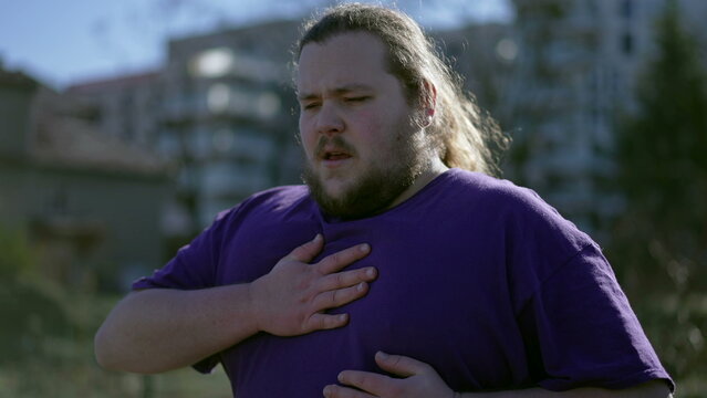 One young overweight man having chest pains standing outdoors. A fat person feeling stressful pain with difficulty in breathing