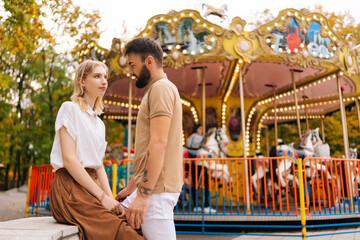 Portrait of romantic young couple in love standing sitting holding hands in amusement park on background of carousel on summer day. Bearded man and pretty woman walking spending time together outdoors