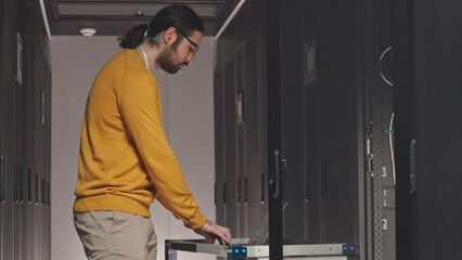 Caucasian IT engineer using laptop while maintaining work of hardware in data center