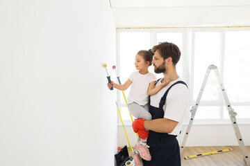 Smiling 5-year-old girl holds a paint roller in her hands. The child helps her father, who holding her, near the wall, in the repair.