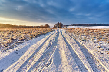 Snowy rural road with traces and footprints on winter riverbank. Frost on grass, cane. February Forest river sunrise. Cold Weather landscape. Low Cloudy sky