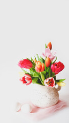 Easter still life with multicolored tulips in a jug and white eggs on white background with copy space. Preparing for Easter. Spring holiday mood. Vertical format