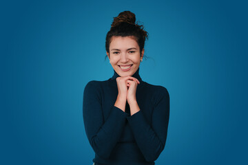 Fototapeta na wymiar Portrait of cute young woman with brunette hair and gentle smile holding hands under chin looking contented at camera isolated over blue background. Attractive