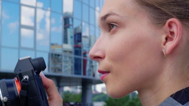 Young woman takes photo while standing next to modern building. Photographer traveler takes pictures of city sights. Positive girl travels in strange town. Side view