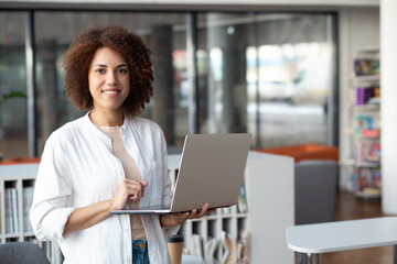 Successful young woman entrepreneur holds laptop, stands in modern office. Portrait of African american businesswoman smiling and looking at the camera