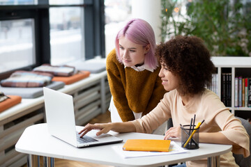 Fototapeta premium Two young female colleagues working on laptop in office. Modern business women entrepreneurs discuss a project, chat, smile. Teamwork collaboration, achieving success together
