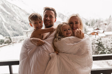 Family standing on balcony covered in blanket in early morning enjoying good winter day in Alps.