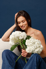 A happy fragile girl in blue jeans and a beige corset sits on a chair and holds a bouquet of hydrangeas in her hands. Blue background.