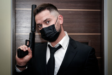 A man of European appearance in a black suit with a mask on his face and with a pistol with a...
