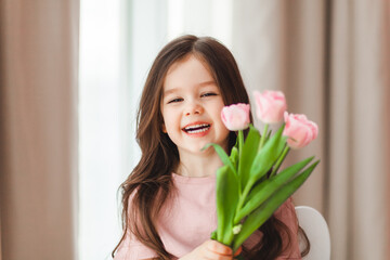 Portrait of a little girl with long dark hair close-up. The baby hugs a bouquet of fresh, delicate pink tulips. A gift for the holiday, spring time.