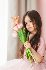 Portrait of a little girl with long dark hair close-up. The baby hugs a bouquet of fresh, delicate pink tulips. A gift for the holiday, spring time.