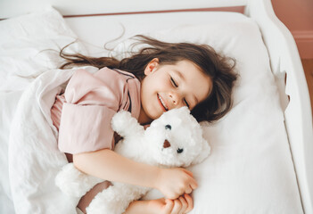 a little dark-haired girl is sleeping in bed, hugging a teddy bear under a white blanket.the child is in bed in the morning.