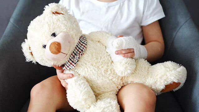 Little girl with broken finger holds teddy bear with a bandaged paw at the doctor's appointment in the hospital
